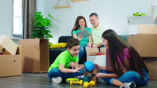 Coppia attraente e i loro figli si sono trasferiti in una nuova casa i bambini stanno giocando sui giocattoli mentre i genitori seduti sul divano e guardando sul futuro piano di design del tablet digitale — Video Stock