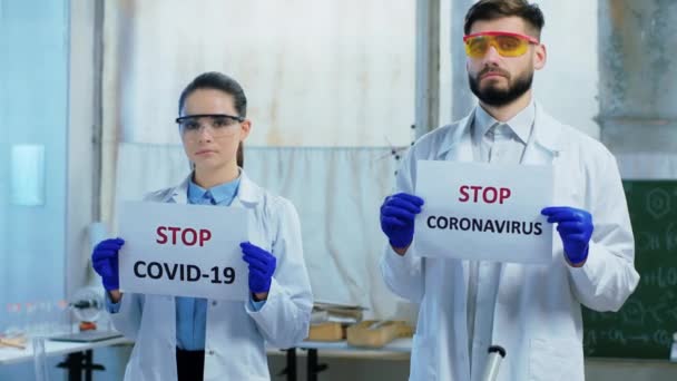 Atractiva mujer y hombre científico en el laboratorio de química frente a la cámara mirando directamente el cartel con Stop Coronavirus. 4k — Vídeos de Stock