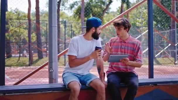 Ragazzi felici in un parco moderno che si godono il tempo insieme usando un tablet per analizzare qualcosa di cui discutono insieme uno dei ragazzi che beve un caffè — Video Stock