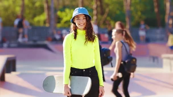 Good looking lady with curly hair in a skate park standing in front of the camera and smiling charismatic while she holding her skateboard — стокове відео