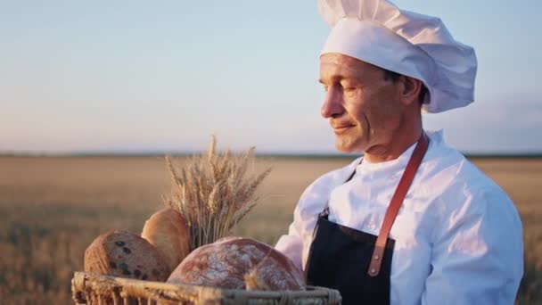 Oude man bakker in een groot tarweveld met een mand vol vers brood kijkt hij serieus recht naar de camera — Stockvideo