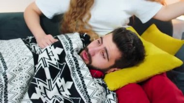 Güzel kahverengi sakallı ve taranmış saçlı hasta bir adam partnerinin kucağında oturuyor, bir battaniyeye sarılmış ve çok hasta hissediyor ve burnunu mendile siliyor. ARRI Alexa 'da çekim