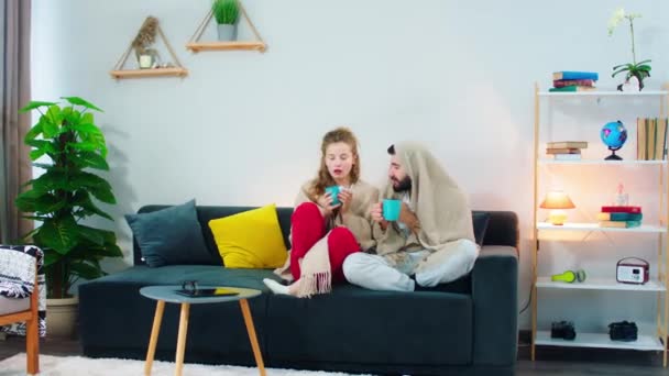 Uma jovem mulher olhando doente e frágil espirros e congelamento no sofá com um homem sentado ao lado dela envolto em um cobertor, ele está olhando confortável — Vídeo de Stock