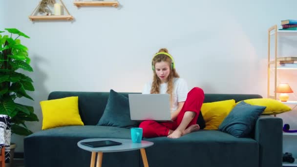 Gyönyörű szőke haj hölgy használ laptop gépelni néhány információt, miközben otthon marad a kanapén dolgozik online visel fejhallgató — Stock videók