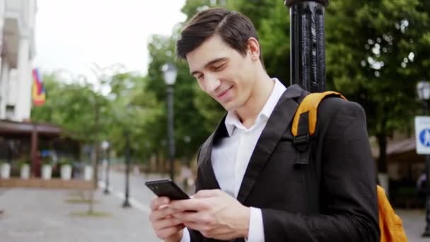Добре виглядає чоловік бізнес виглядає з великою посмішкою, вводячи щось на свій смартфон посеред вулиці — стокове відео