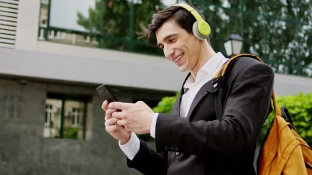 Blízko k fotoaparátu charismatický muž v obleku pomocí telefonu na psaní něco při chůzi po ulici má na sobě zelené sluchátka poslouchat hudbu — Stock video