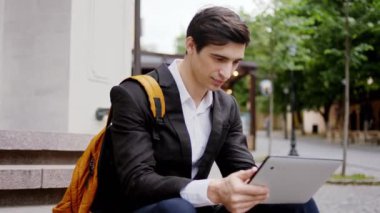 Siyah takım elbiseli konsantre öğrenci elektronik tabletini al ve sokağın ortasında merdivenlerde otururken bir şeyler yazmaya başla.