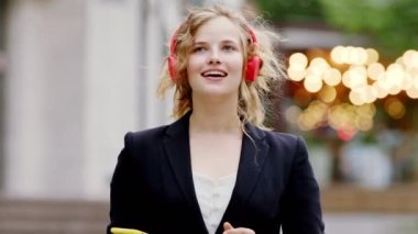 Ranzalı kulaklık takan karizmatik sarışın kadın modern caddenin ortasında mutlu bir şekilde gülümseyen bir telefondan müzik dinliyor. ARRI Alexa Mini 'de çekilen