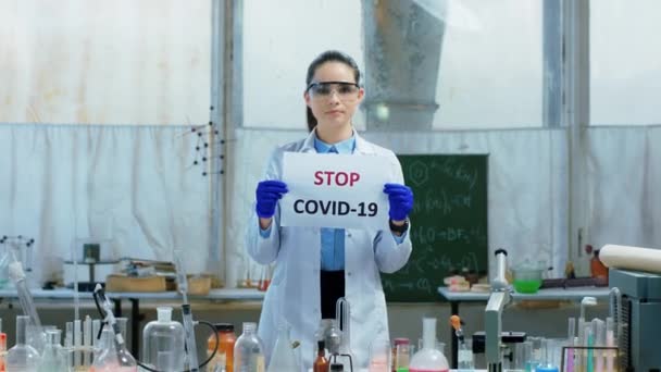 Zbliżenie do kamery młoda kobieta naukowiec w laboratorium chemicznym trzymając plakat z Stop COVID-19 patrzy prosto do kamery — Wideo stockowe