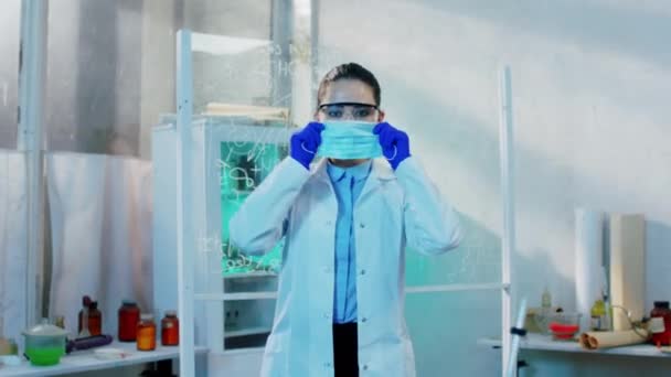 En el laboratorio bioquímico mujer científica de buen aspecto con uniforme protector obtener la máscara protectora y el uso en la cara y posando delante de la cámara — Vídeos de Stock