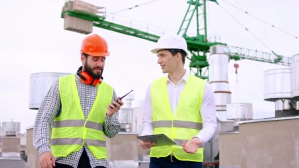 Два парня в защитных шлемах один бизнесмен и один парень идут по крыше и анализируют план строительства с помощью цифрового планшета — стоковое видео