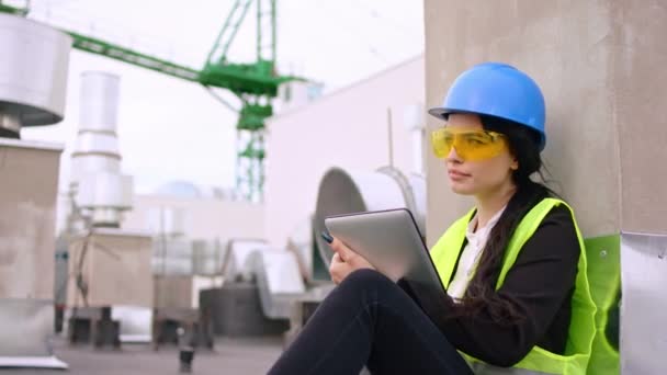 Молодая ассистентка инженера на верхней части строительной площадки садится на пол в защитном шлеме и очках, у нее в руках цифровой планшет — стоковое видео