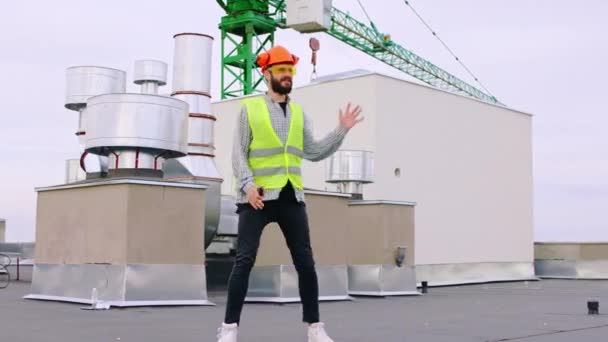 Χορός όμορφος άντρας μπροστά από την κάμερα στην κορυφή του εργοταξίου φοράει κράνος και εξοπλισμό ασφαλείας — Αρχείο Βίντεο