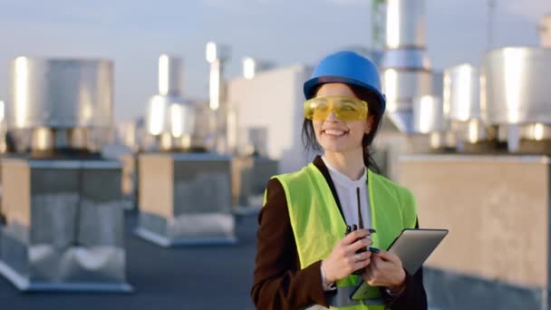 Fiatal nő mérnök, nagy mosollyal, sárga építőszemüveget és biztonsági sisakot visel, a kamera előtt áll, és egy térképet tart egy tervvel és nagy mosollyal. — Stock videók