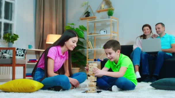 En la sala de estar chica bonita y su hermano pequeño jugando juntos en el juego de madera, mientras que sus padres pasan tiempo en el ordenador portátil mientras están sentados en el sofá — Vídeos de Stock