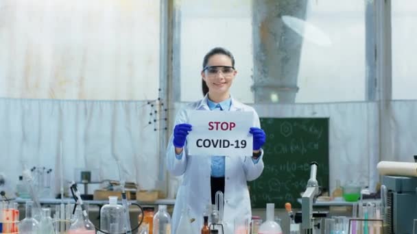 Di laboratorium biokimia tersenyum besar wanita cantik ilmuwan memegang poster Stop COVID-19 melihat langsung ke kamera — Stok Video