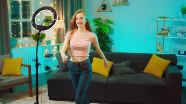 집 거실에 있는 예쁜 여자가 소셜 미디어 계정을 위한 댄스 영화를 찍고 있습니다. 그녀의 스마트폰에서 라이트 링을 이용해 비디오를 녹화하고 있습니다. — 비디오