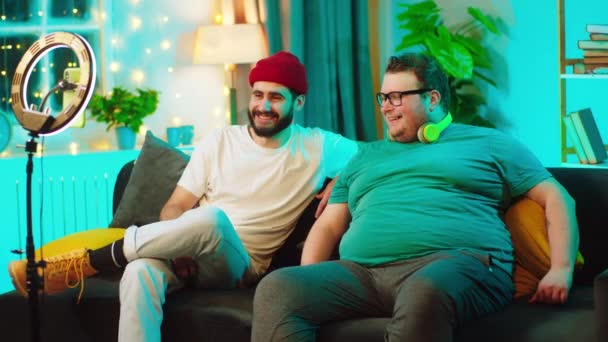 Auf dem Sofa unterhalten sich zwei charismatische Männer, von denen einer fettleibig ist, im Livestream von einem Social-Media-Account, der vom Smartphone aufgezeichnet wird. — Stockvideo