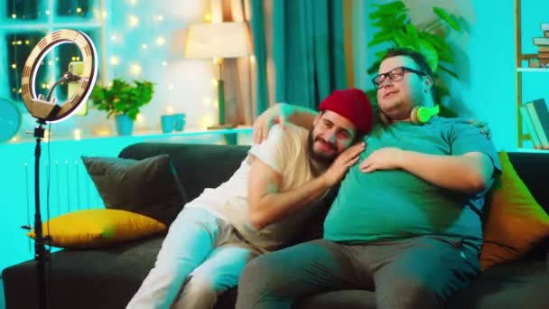 Un couple gay influenceurs passer un agréable moment ensemble sur le canapé à la maison ils enregistrent film drôle pour le compte de médias sociaux en utilisant un smartphone pour enregistrer — Video