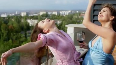 Pijamalı güzel bayanlar çatı katının çatısında temiz havanın tadını çıkarıyorlar mutlu bir şekilde dans ediyorlar büyük bir gülümseme ve bir kadeh şarapla şerefe. 4k