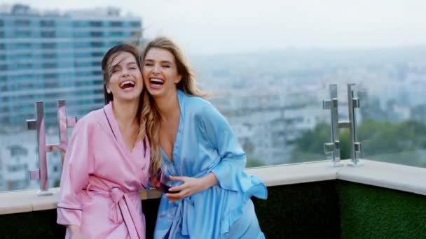 Perfeito sorrindo duas belas senhoras de pijama no topo do terraço com vista para a cidade posando na frente da câmera e segurando copos de vinho. Tiro em ARRI Alexa — Vídeo de Stock