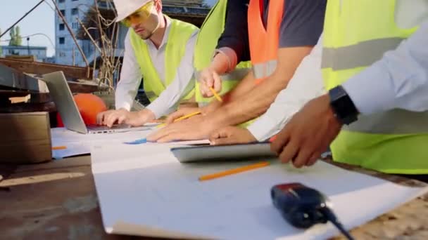 Grupo de diversos especialistas no canteiro de obras usando tablet digital e grande plano de construção local analisando e fazer algumas mudanças que vestem uniforme de proteção e capacetes de segurança — Vídeo de Stock