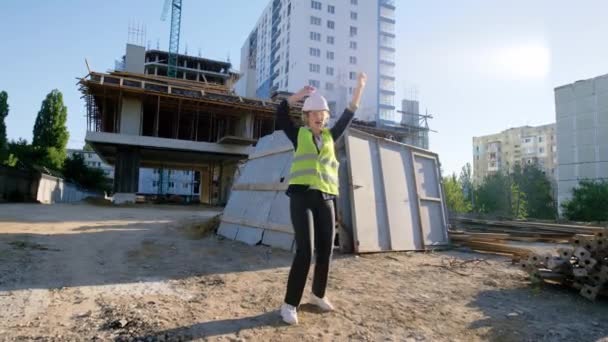 Tanzende junge Ingenieurin mit Schutzhelm und Schutzbrille auf der Baustelle, sie lächelt groß — Stockvideo