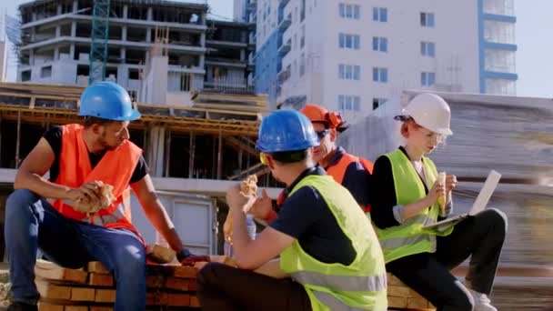 建設現場の多様な専門家のグループは、休憩時間を持っている彼らはいくつかのサンドイッチを食べ、安全制服を着て議論を持っている — ストック動画