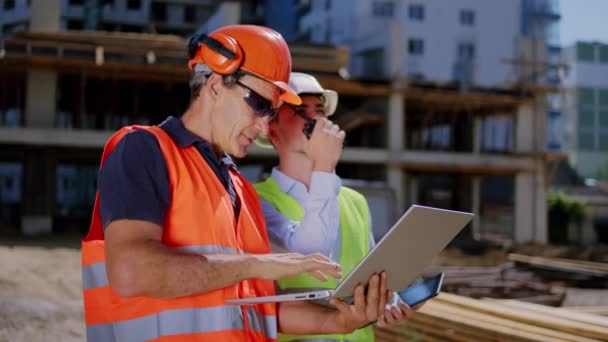 Acercamiento al hombre constructor de la cámara y arquitecto utilizando el ordenador portátil y la ración para hablar con otros trabajadores de la obra de construcción que analizan el plan de construcción que llevan cascos de seguridad y — Vídeo de stock