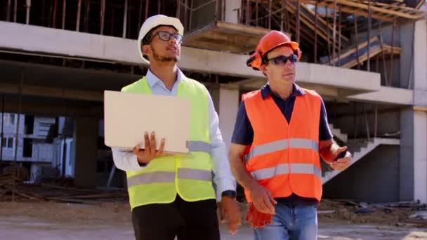 Charismatischer afroamerikanischer Ingenieur läuft mit Laptop neben dem Polier auf der Baustelle und analysiert den zukünftigen Bauplan in Sicherheitsausrüstung — Stockvideo