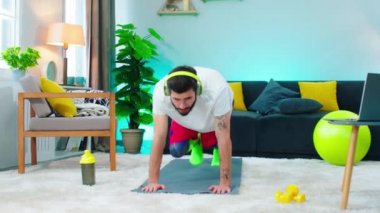 Kahverengi sakallı ve saçlı bir adam, spor kıyafetleri giyiyor ve gri bir Matt 'in üzerinde dağa tırmanma egzersizi yapıyor, dizüstü bilgisayarına bakmak için duruyor. 4k