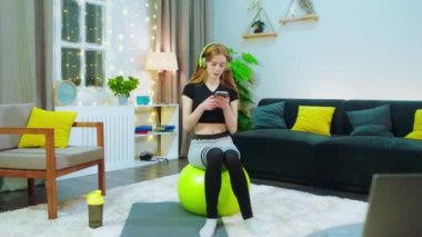 Sarı bir yoga topunun üzerinde oturan bir kız sarı bir kulaklık takıyor, antrenman yapmak üzere ve müzik dinliyor, sarı koordine şeyler olan bir odada.