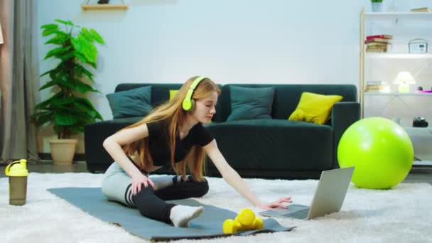 쿨 해 보이는 여자가 파란색 매트 위 바닥에 다리를 뻗고 있습니다. 노란색 헤드셋을 쓰고 있고 회색 레 깅스를입고 있고, 옆에 는 컴퓨터가 있어서 운동을 도와 줍니다. — 비디오