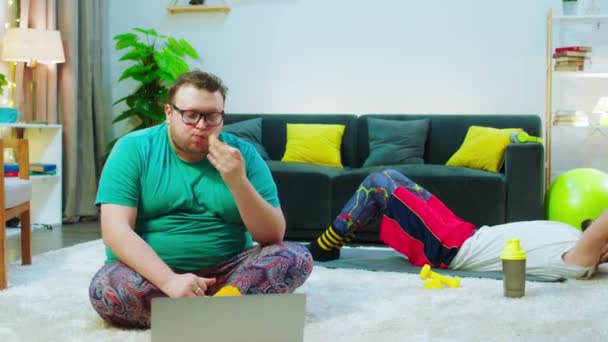 덩치큰 뚱보가 햄버거를 먹으면서 노트북 위의 영화를 보고 있는 동안 바닥에 앉아 있는 동안 다른 덩치큰 남자는 6 팩을 받기 위해 와치를 하고 있습니다. ARRI 알렉사 주사 — 비디오