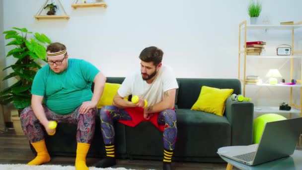 Zu Hause zwei Freunde einer von ihnen sind fettleibig beim gemeinsamen Workout, während sie auf dem Sofa sitzen sie mit Gewichten Muskeln machen — Stockvideo