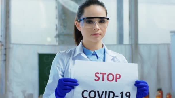 W koncepcji pandemii farmaceutka z laboratorium chemicznego trzymająca plakat Stop COVID-19 przed kamerą. Strzał na ARRI Alexa Mini. — Wideo stockowe