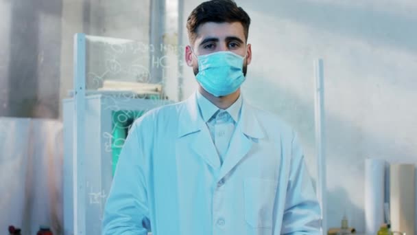 コロナウイルスの隔離で保護マスクでカメラの前にポーズ医薬品研究所のいい顔の男薬剤師 — ストック動画