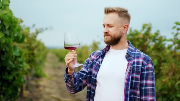 Charyzmatyczny mężczyzna przed kamerą bierze kieliszek wina i smakuje na środku winnicy, którą pije z przyjemnością. — Wideo stockowe