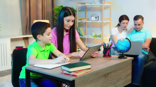 In de moderne ruime woonkamer zus en kleine broer samen helpen elkaar doen huiswerk, terwijl ouders op de bank genieten van de avond tijd — Stockvideo