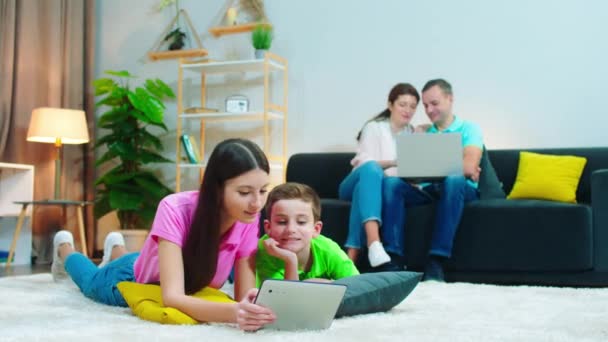 Feliz família grande passar um bom tempo juntos na sala de estar pais sentados no sofá e usando laptop para assistir a algo juntos, enquanto as crianças usando o tablet digital para jogar algum jogo. — Vídeo de Stock