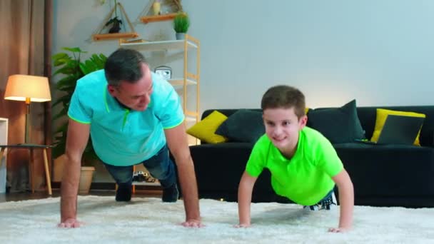 Lachende grote vader en zoon doen thuis training op de vloer in de woonkamer vader laten zien hoe te schrijven om de push-ups te doen — Stockvideo