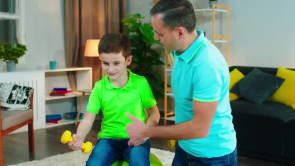 Άσκηση του χρόνου για τον μπαμπά και χαριτωμένο γιο του στο σαλόνι χρησιμοποιώντας βάρη για να κάνουν τους μυς μαζί εργάζονται την ίδια στιγμή — Αρχείο Βίντεο