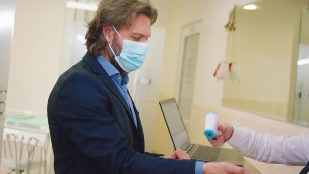 Асистент клініки на прийомі перевіряє температуру пацієнта, в якому вони носять захисну маску при пандемії коронавірусу, а потім людина робить знак на історію відвідування — стокове відео