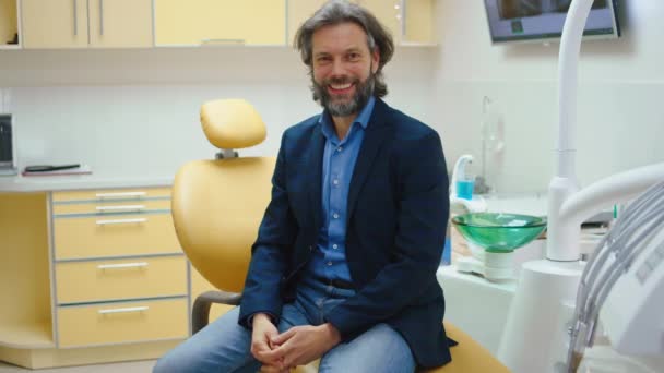 Hombre carismático paciente en la clínica dental tomar un asiento en la silla del dentista y sonriendo grande con los dientes perfectos que muestra grande como a la cámara — Vídeos de Stock
