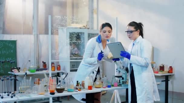 Deux collègues scientifiques femmes dans le laboratoire pharmaceutique analysant la formule chimique à l'aide de la tablette numérique pour écrire quelques notes — Video