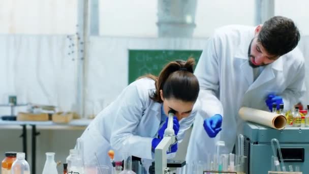 Ученый мужчина и женщина рассматривают микроскоп вместе со своим коллегой, обсуждая в лаборатории химии концепцию научно-испытательной лабораторной промышленности — стоковое видео