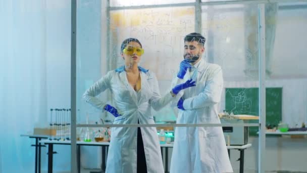 Femeia farmacistă atractivă și bărbatul ei coleg care lucrează împreună în laboratorul farmaceutic analizează formula chimică din placa transparentă — Videoclip de stoc
