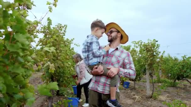 Happy and smiling large farmer man and his smiling cute lovely boy in the middle of the vineyard they spending time together in a autumn day background other seasonal workers collecting the grapes — Stockvideo