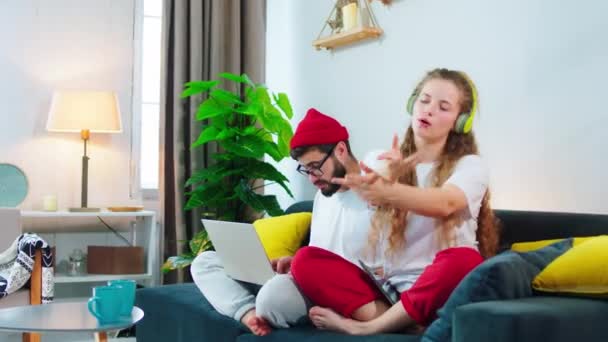 At home young couple very attractive and funny spending the time together lady singing and dancing while sitting on the sofa beside of her boyfriend while him is using laptop for work — Stockvideo