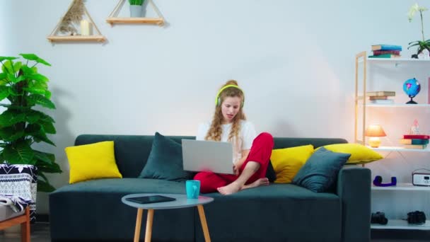 Bella donna con i capelli ricci godendo il tempo a casa mentre fa un po 'di lavoro online lei ascoltando una buona musica dalle cuffie. Girato su ARRI Alexa — Video Stock